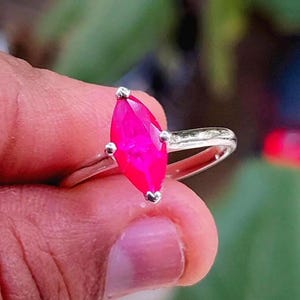 Puede incluir: Un anillo de plata con una gema fucsia brillante en forma de marquesa. La gema está engastada con cuatro garras, y la banda es de plata pulida. El anillo se encuentra sobre un fondo verde borroso.