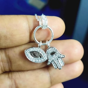 May include: Silver pendant necklace featuring an eye and a hand charm, both encrusted with small, clear gemstones. The eye charm has a dark brown center. The necklace is held by a hand against a blue background.
