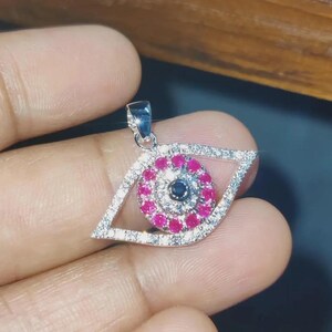 May include: A silver-coloured eye-shaped pendant. The iris is a circle of pink gemstones with a dark blue centre. The eye's outline is formed by small, clear gemstones.