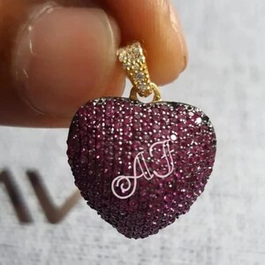 May include: A heart-shaped pendant with a gold bail, set with small, dark pink stones. The pendant has the initials "AF" in white, cursive script. The bail is decorated with small, clear stones.