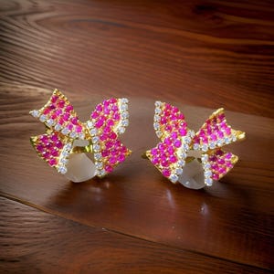 May include: A pair of gold-coloured bow-shaped earrings. The bows are encrusted with small, bright pink gemstones and outlined with clear, sparkling stones. Each earring has a large, pearly white stone at the centre.