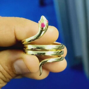 May include: A gold-colored snake ring with a coiled design. The snake's head is adorned with two small, red gemstones. The ring wraps around the finger, creating a unique and eye-catching accessory.