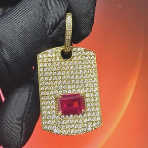Könnte beinhalten: Goldfarbener Anhänger in rechteckiger Form, besetzt mit kleinen, klaren Edelsteinen. In der Mitte befindet sich ein rechteckiger, tiefroter Edelstein. Der Anhänger hat oben eine Schlaufe, die ebenfalls mit klaren Edelsteinen verziert ist.
