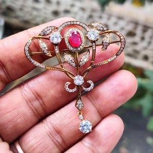 Könnte beinhalten: Goldfarbene Brosche mit einem ovalen Rubin-Mittelstein, umgeben von kleinen Diamanten. Die Brosche hat ein florales Design mit zusätzlichen Diamantakzenten und einem hängenden Diamantanhänger.
