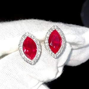 Op de afbeelding: Een paar marquise-vormige oorbellen met een levendig rode edelsteen in het midden, omringd door een halo van kleine, sprankelende witte stenen. De oorbellen zijn gezet in een zilverkleurig metaal, wat een luxueus en opvallend ontwerp creëert.