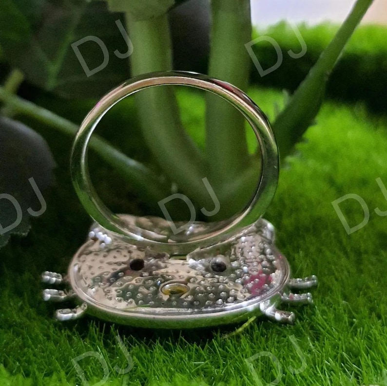 May include: A silver ring with a frog-shaped base. The ring band is a simple, polished silver. The frog base has a textured surface with small details, including eyes and feet. The ring is set against a backdrop of green foliage and grass.
