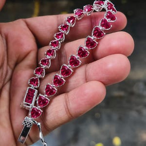 Puede incluir: Una pulsera plateada con piedras preciosas rojas en forma de corazón. La pulsera está compuesta por una serie de piedras en forma de corazón unidas por una cadena plateada. El cierre también es plateado y rectangular.