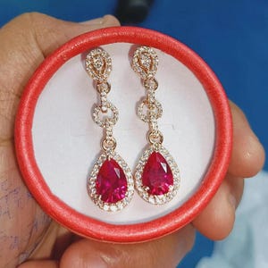 May include: A pair of gold-toned drop earrings with a teardrop-shaped, ruby-red center stone. The earrings are surrounded by a halo of small, clear stones and are presented in a red jewelry box.