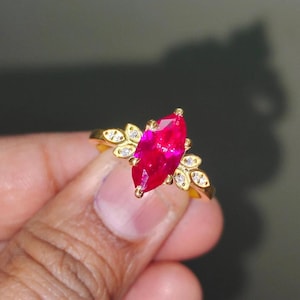 Peut inclure: Une bague en or avec une grande pierre précieuse de rubis de forme marquise rose vif entourée de petites pierres précieuses claires.