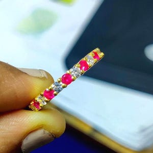 May include: A gold ring with alternating red and clear gemstones. The ring is held between fingers, showcasing the vibrant colors of the gemstones. The ring is a piece of jewelry.