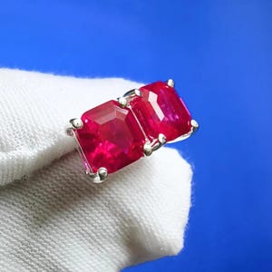 Könnte beinhalten: Ein Paar quadratisch geschliffene, leuchtend rote Edelsteinohrringe, gefasst in Silber. Die Edelsteine werden von kleinen Zinken gehalten, und die Ohrringe werden vor einem weißen Stoff und einem blauen Hintergrund präsentiert.