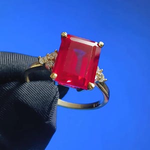 May include: A gold ring featuring a large, red, emerald-cut gemstone and smaller clear gemstones on the band.