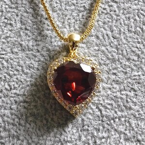 Op de afbeelding: Een gouden ketting met een hartvormige hanger, bezet met een grote rode edelsteen omringd door kleinere heldere edelstenen.
