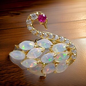 May include: A gold swan brooch featuring a pink gemstone at the centre of the swan's head. The swan is decorated with white opals and clear cubic zirconia stones.