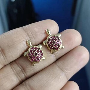 Peut inclure: Deux boucles d'oreilles dorées en forme de tortue. Chaque boucle d'oreille présente une carapace recouverte de petites pierres précieuses rondes et rouges. Les boucles d'oreilles sont présentées sur un fond flou.