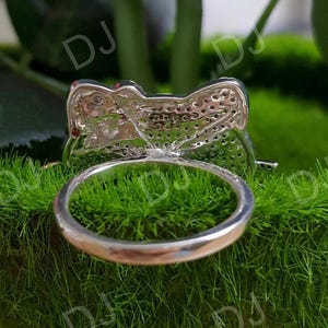 May include: A silver ring with a bow-shaped design, set with small, sparkling stones. The ring is shown against a backdrop of green moss, highlighting its intricate details and reflective surface. The ring's band is smooth and polished.