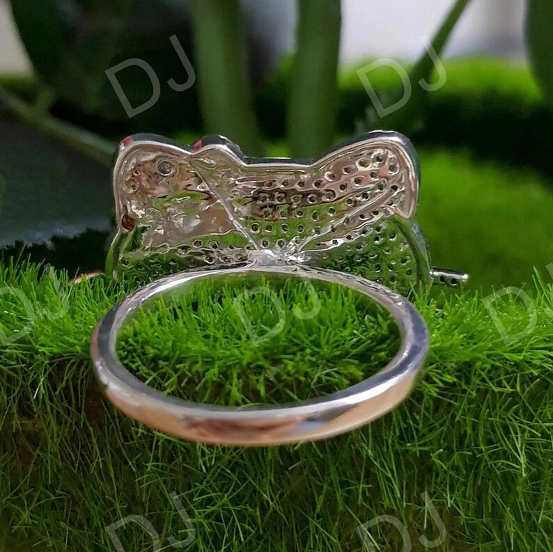 May include: A silver ring with a bow-shaped design, set with small, sparkling stones. The ring is shown against a backdrop of green moss, highlighting its intricate details and reflective surface. The ring's band is smooth and polished.