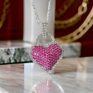 Peut inclure: Un collier en argent avec un pendentif en forme de cœur. Le pendentif est recouvert de petites pierres précieuses roses et entouré de petites pierres précieuses blanches.