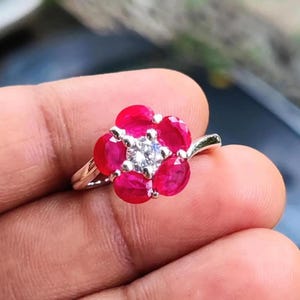 Può includere: Un anello d'argento con un motivo floreale. Il fiore è composto da cinque pietre preziose rosse ovali vivaci che circondano un gruppo di pietre preziose più piccole e trasparenti. L'anello è tenuto in mano, evidenziando i dettagli.