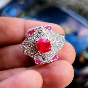 May include: A silver ring featuring a large, square, red gemstone. The ring has an intricate design with small, clear stones and pink accents. The ring is held in a hand, showing the detailed craftsmanship.