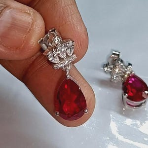 May include: A pair of silver drop earrings. Each earring has a teardrop-shaped, deep red gemstone hanging from a cluster of small, clear stones. The earrings are held up by a silver post.