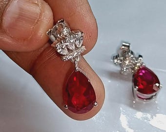 May include: A pair of silver drop earrings. Each earring has a teardrop-shaped, deep red gemstone hanging from a cluster of small, clear stones. The earrings are held up by a silver post.