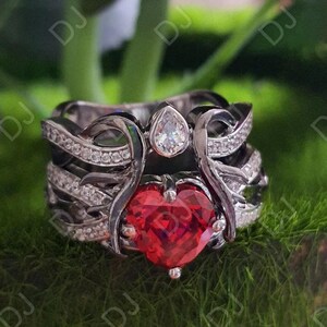 Può includere: Un set di anelli a tre pezzi con una gemma rossa a forma di cuore e una gemma bianca a forma di pera. Gli anelli hanno una finitura in metallo scuro e sono impreziositi da piccole gemme trasparenti. Il set è esposto su muschio verde.