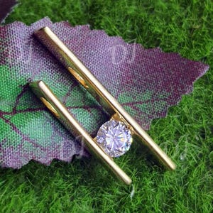 Puede incluir: Un pasador de corbata dorado con una gema redonda y transparente en el centro. El pasador de corbata descansa sobre una tela estampada en morado y verde, sobre un fondo de hierba verde.
