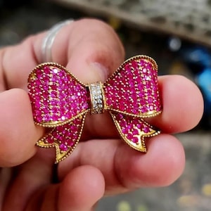 May include: A gold-coloured bow brooch, adorned with numerous small, bright pink gemstones. The centre of the bow has a band of clear, sparkling stones. The brooch is held in a hand, highlighting its detailed design and craftsmanship.
