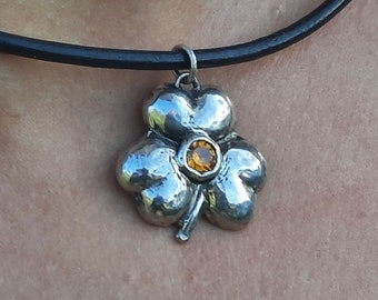 Op de afbeelding: Een zilveren vierbladige klaverhanger met een amberkleurige edelsteen in het midden, hangend aan een zwart leren koord.