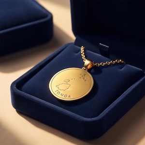 May include: A gold-toned circular pendant necklace featuring an engraved map outline of Tonga and the text TONGA. The pendant is attached to a delicate gold-toned chain and rests inside a dark blue velvet-lined jewellery box with a matching lid.