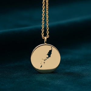 May include: Gold pendant necklace with a circular charm showing a detailed landmass cutout. The necklace has a fine chain and is set against a dark teal backdrop. The pendant is approximately 2.5 cm in diameter.