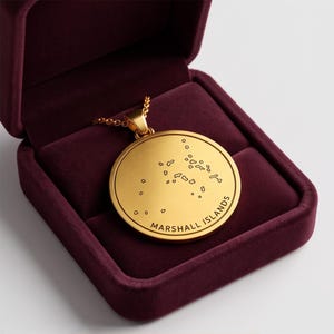 Puede incluir: Un collar con colgante circular de tono dorado que presenta un mapa grabado de las Islas Marshall y el texto "MARSHALL ISLANDS" a lo largo del borde inferior. El colgante se muestra dentro de un joyero forrado en terciopelo burdeos.
