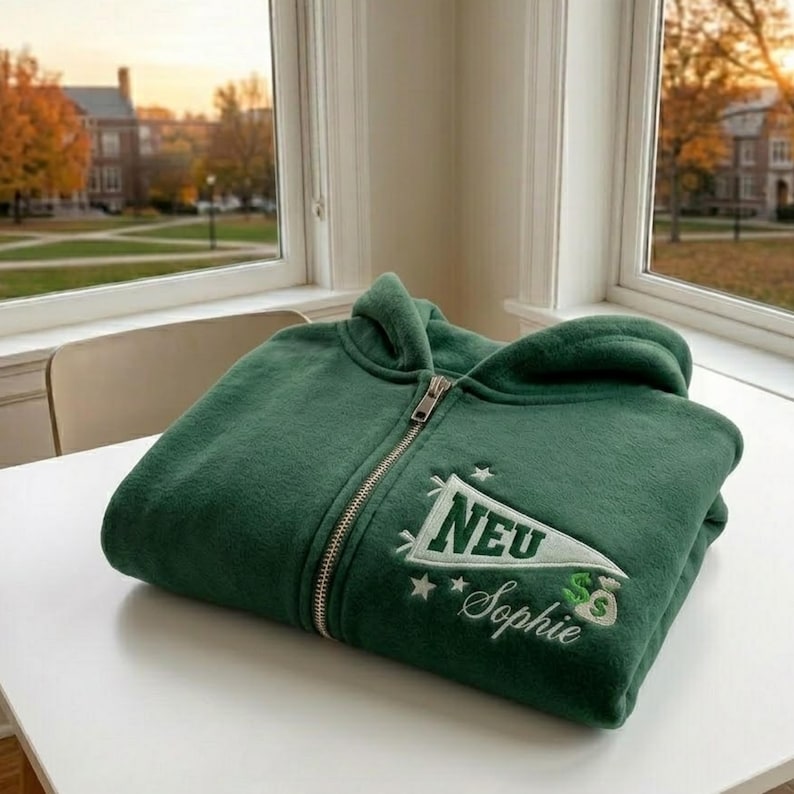 May include: A folded, forest green zip-up hoodie with a white and green embroidered pennant design. The pennant reads "NEU" and "Sophie" with dollar signs. The hoodie is on a white surface near a window.