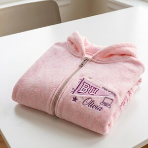 May include: A folded light pink fleece zip-up hoodie. The hoodie features a silver zipper and a purple embroidered design with the letters "BU", a star, a laptop, the words "Olivia" and "Class of 2026".