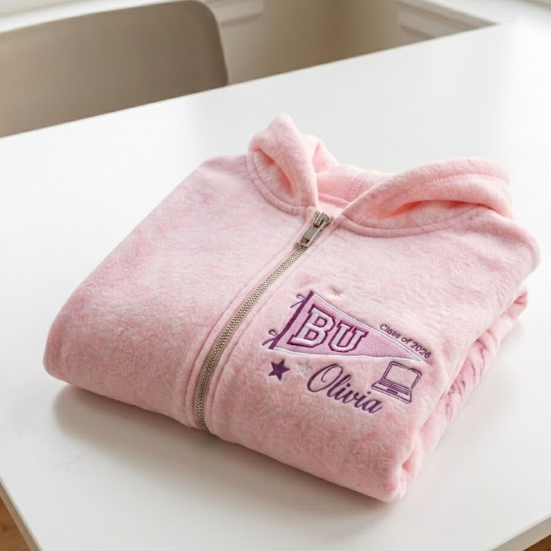 May include: A folded light pink fleece zip-up hoodie. The hoodie features a silver zipper and a purple embroidered design with the letters "BU", a star, a laptop, the words "Olivia" and "Class of 2026".