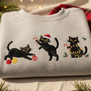May include: A light gray sweatshirt featuring embroidered black cats with yellow eyes, decorated for Christmas. One cat plays with ornaments, another holds a candy cane, and the third is adorned with lights. Festive holiday apparel.