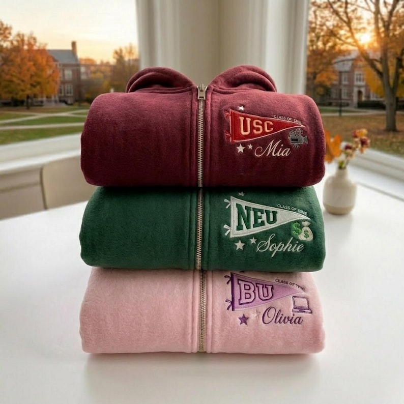 May include: Three stacked fleece zip-up hoodies in burgundy, green, and pink. Each hoodie features a school pennant design with embroidered text: "USC Mia", "NEU Sophie", and "BU Olivia". The hoodies are stacked on a white surface.