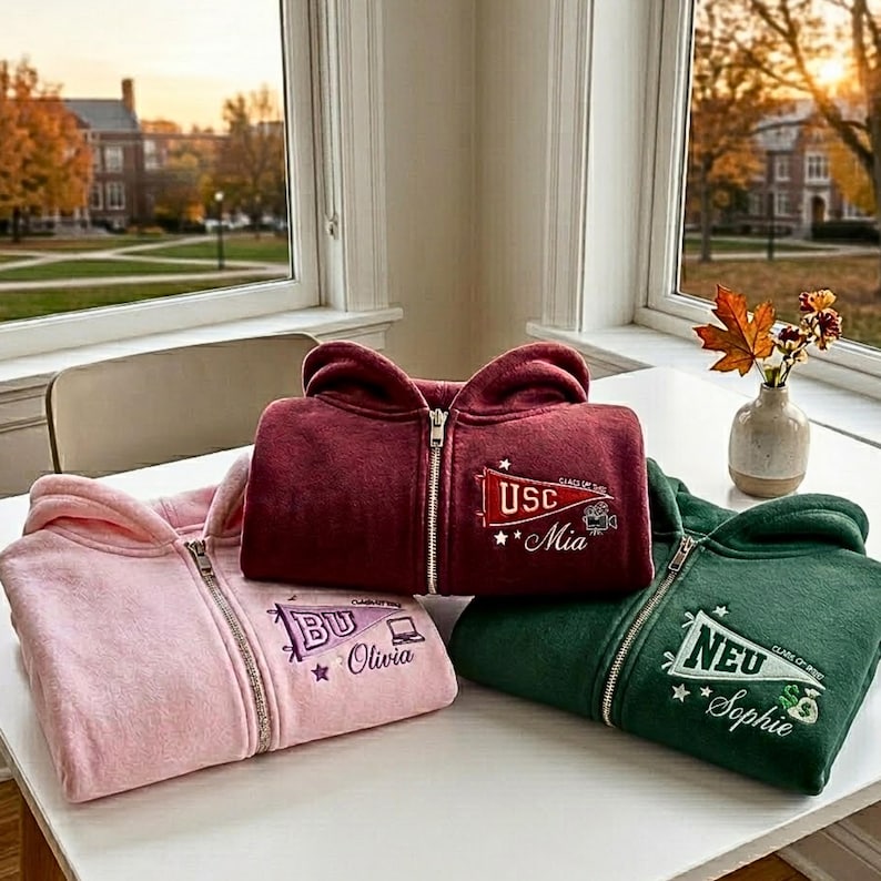 May include: Four zip-up hoodies in burgundy, pink, and green are displayed on a white surface. Each hoodie features embroidered text and graphics, including school names and student names. A small vase with fall foliage sits nearby.