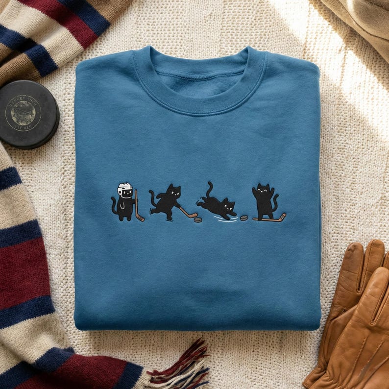 May include: Teal blue sweatshirt featuring a playful graphic of black cats playing hockey. The cats are depicted in various hockey-related poses, with hockey sticks and a puck. The sweatshirt is made of a soft material.