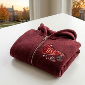 May include: A folded maroon zip-up hoodie with a hood and a silver zipper. The hoodie features an embroidered patch with the text "USC", "Mia", and "Class of 2026". The hoodie is made of a soft, plush material.