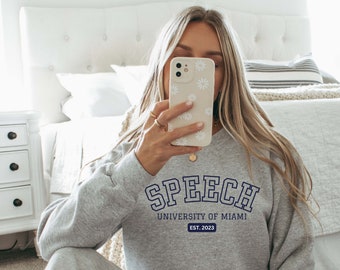 Puede incluir: Sudadera gris con el texto "SPEECH UNIVERSITY OF MIAMI EST. 2023" en azul.