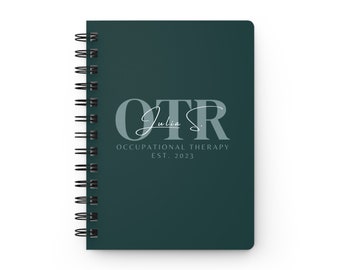May include: A dark green spiral bound notebook with the text "OTR Julia S. OCCUPATIONAL THERAPY EST. 2023" in white and light gray lettering.