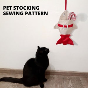 Puede incluir: Una media de color beige con forma de pez con ribete rojo y cinturón rojo cuelga de una pared blanca. Un gato negro se sienta en un suelo de madera mirando hacia arriba a la media. El texto "PET STOCKING SEWING PATTERN" está en la parte superior de la imagen.