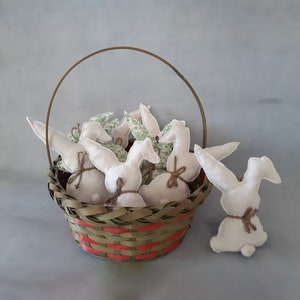 Op de afbeelding: Een rieten mand gevuld met witte stoffen konijnen met groene bloemenaccenten. De mand heeft een rood en groen geweven patroon.