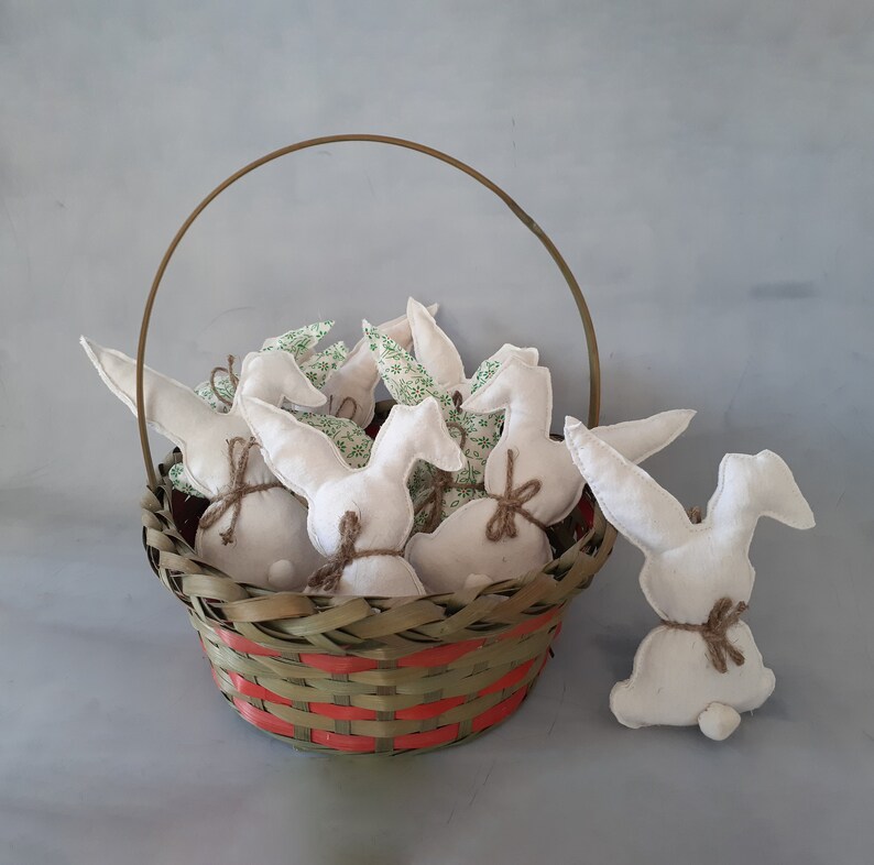Op de afbeelding: Een rieten mand gevuld met witte stoffen konijnen met groene bloemenaccenten. De mand heeft een rood en groen geweven patroon.