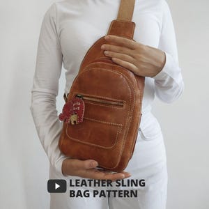 Könnte beinhalten: Eine braune Leder-Umhängetasche mit einer Vordertasche und einem Reißverschluss. Die Tasche hat einen beigefarbenen Riemen und einen dekorativen roten Elefantenanhänger mit der Aufschrift "with love". Der Text "LEATHER SLING BAG PATTERN" befindet sich unten.