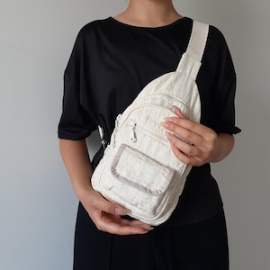 May include: Cream-colored sling bag with a textured pattern. Features a zippered main compartment, a smaller front pocket, and an adjustable strap. The bag is held against a person wearing a black top.