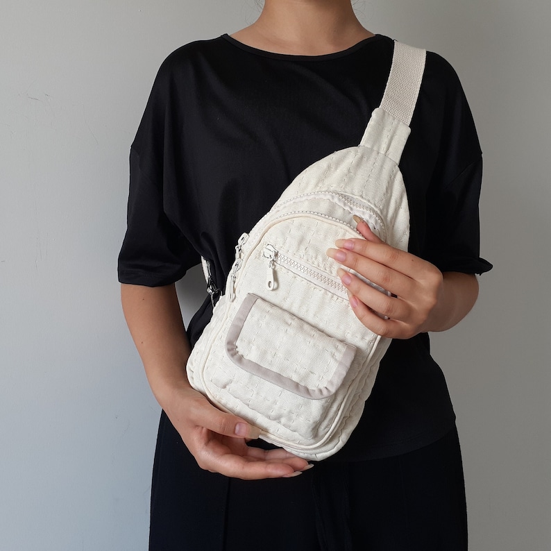 May include: Cream-colored sling bag with a textured pattern. Features a zippered main compartment, a smaller front pocket, and an adjustable strap. The bag is held against a person wearing a black top.