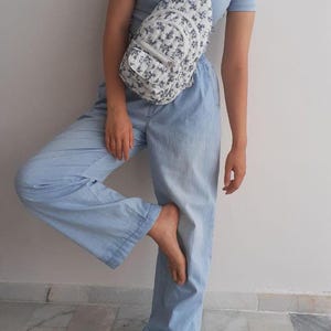 May include: A white sling bag with a blue floral pattern is worn over the shoulder. The bag has a small front pocket and a matching strap. The person is wearing a light blue top and light blue pants.